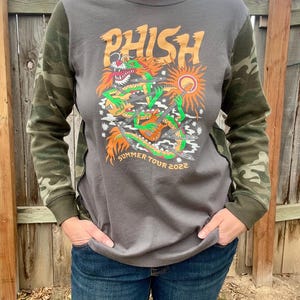 May include: A gray long-sleeve shirt with a camouflage print on the sleeves. The shirt features a graphic of a cartoon dragon playing a guitar with the word "Phish" above it and the text "Summer Tour 2022" below it.