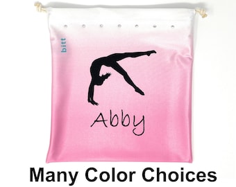 May include: Pink and white drawstring bag with a silhouette of a gymnast doing a back handspring. The bag has the word "bitt" in blue text and the name "Abby" in black text.