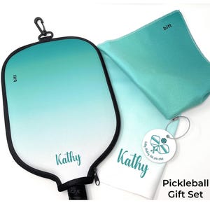 Può includere: Una racchetta da pickleball color verde acqua e bianco sfumato con un manico nero e un asciugamano abbinato color verde acqua e bianco sfumato con il nome "Kathy" stampato su di esso. L'asciugamano ha anche un piccolo cerchio bianco con una grafica di pickleball e il testo "Kathy" stampato su di esso. Il testo "Pickleball Gift Set" è stampato sotto l'asciugamano.