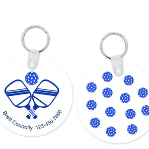 Pu&ograve; includere: Portachiavi rotondo bianco con un design pickleball blu. Un lato presenta racchette incrociate e una palla, con testo. L'altro lato ha un motivo di palline da pickleball blu e bianche. Include un portachiavi in metallo.