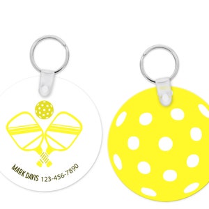 Pu&ograve; includere: Un portachiavi rotondo con una clip in plastica bianca e un anello in metallo argentato. Un lato presenta un grafico di pickleball giallo e racchette incrociate con il testo "MARK DAVIS 123-456-7890". L'altro lato &egrave; un cerchio giallo con punti bianchi.
