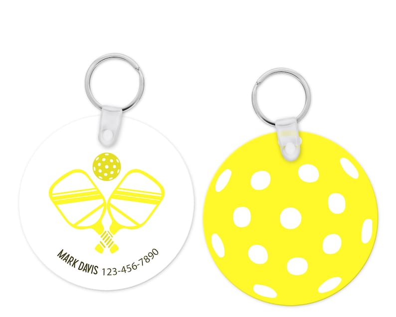 Pu&ograve; includere: Un portachiavi rotondo con una clip in plastica bianca e un anello in metallo argentato. Un lato presenta un grafico di pickleball giallo e racchette incrociate con il testo "MARK DAVIS 123-456-7890". L'altro lato &egrave; un cerchio giallo con punti bianchi.