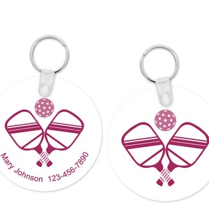 Pu&ograve; includere: Due portachiavi rotondi bianchi con un portachiavi argentato. Ogni portachiavi presenta un design pickleball magenta con racchette incrociate e una palla. Un portachiavi ha il nome "Mary Johnson" e un numero di telefono.