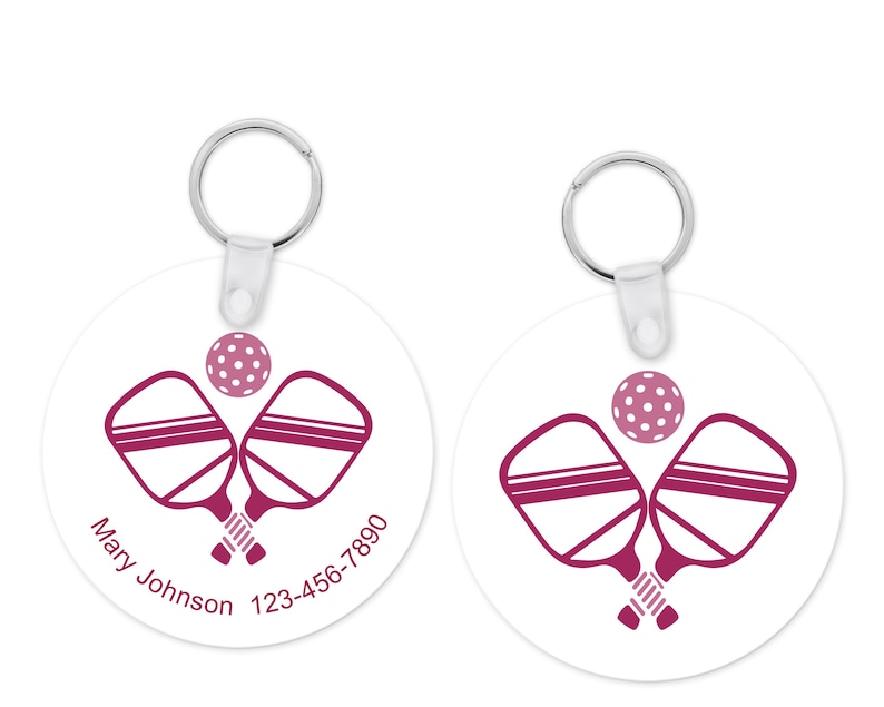 Pu&ograve; includere: Due portachiavi rotondi bianchi con un portachiavi argentato. Ogni portachiavi presenta un design pickleball magenta con racchette incrociate e una palla. Un portachiavi ha il nome "Mary Johnson" e un numero di telefono.