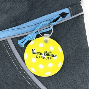 Pu&ograve; includere: Un'etichetta per bagagli gialla e circolare con pois bianchi e il nome "Karen Bittner" e un numero di telefono stampati in nero. L'etichetta &egrave; attaccata a un cordino blu e a un portachiavi argentato, su uno zaino grigio.