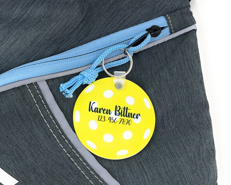 Pu&ograve; includere: Un'etichetta per bagagli gialla e circolare con pois bianchi e il nome "Karen Bittner" e un numero di telefono stampati in nero. L'etichetta &egrave; attaccata a un cordino blu e a un portachiavi argentato, su uno zaino grigio.