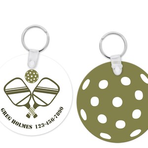 Pu&ograve; includere: Un portachiavi rotondo con un tema pickleball. Un lato mostra racchette da pickleball incrociate e una palla, con il testo "GREG HOLMES 123-456-7890". L'altro lato &egrave; verde oliva con cerchi bianchi. Include un portachiavi argentato.