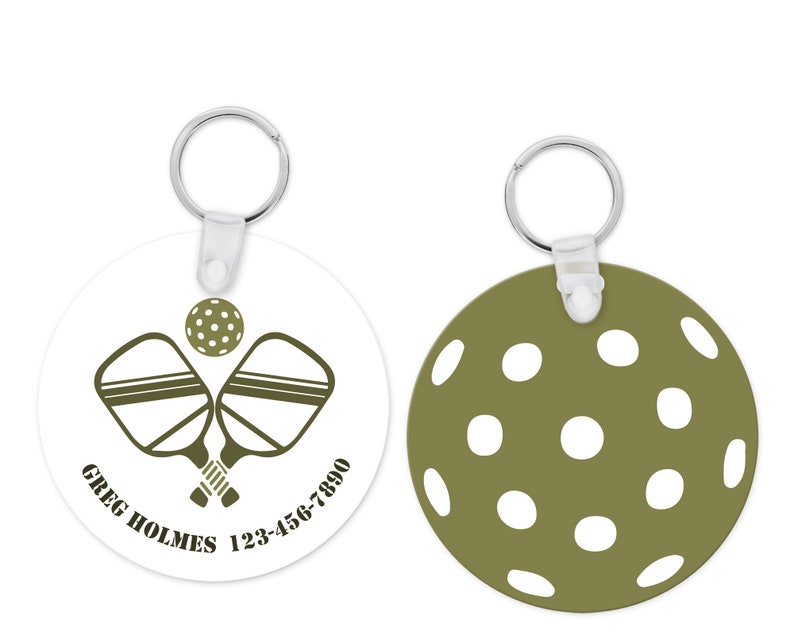 Pu&ograve; includere: Un portachiavi rotondo con un tema pickleball. Un lato mostra racchette da pickleball incrociate e una palla, con il testo "GREG HOLMES 123-456-7890". L'altro lato &egrave; verde oliva con cerchi bianchi. Include un portachiavi argentato.