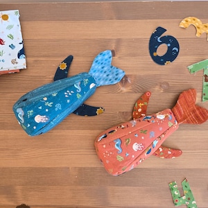 Puede incluir: Dos estuches con cremallera con forma de ballena en azul y coral, adornados con diseños de criaturas marinas. Los estuches miden aproximadamente 20 cm x 10 cm x 5 cm. También aparece el texto "WHALE ZIPPER POUCH" y "Sew a Story".