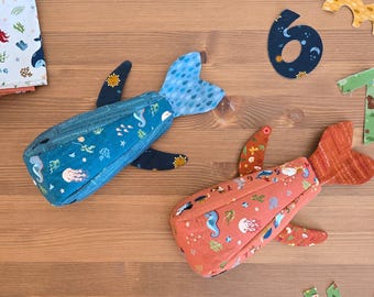 Può includere: Due astucci con cerniera a forma di balena, blu e corallo, decorati con motivi di creature marine. Gli astucci misurano circa 20 cm x 10 cm x 5 cm. Il testo "WHALE ZIPPER POUCH" e "Sew a Story" è presente.