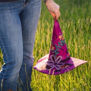 Könnte beinhalten: Ein gesteppter Hexenhut in Lila, verziert mit floralen Applikationen. Der Hut wird vor dem Hintergrund von hohem, grünem Gras und einer Jeans gehalten. Der Text "QUILTED WITCH'S HAT" ist oben im Bild zu sehen.