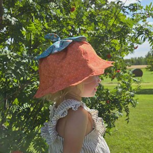 Könnte beinhalten: Ein Kind trägt einen orangefarbenen, blütenblattförmigen Hut mit weißen Punkten und einem blauen Akzent. Das Bild enthält den Text "PETAL PLAY HAT" und "Sew A Story". Der Hut ist ein Schnittmuster für Kinder.