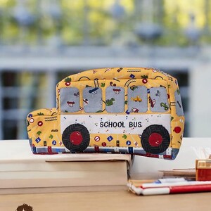 Könnte beinhalten: Ein gelbes Federmäppchen in Form eines Schulbusses mit schwarzen und roten Rädern, blauen Fenstern und dem Schriftzug "SCHOOL BUS" an der Seite. Das Federmäppchen ist mit verschiedenen Fahrzeug- und Straßenmustern verziert. Fertige Größe: 29.2 cm x 6.4 cm x 14 cm.