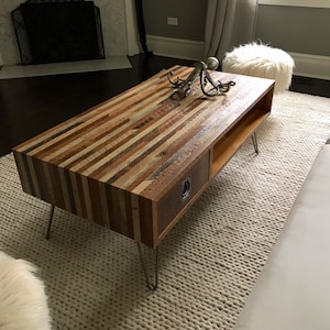 May include: A rustic wooden coffee table with a drawer and a shelf. The table has a unique design with different colored wood strips. The table is on a white rug with a white furry ottoman.