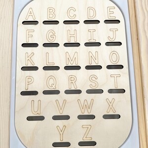 May include: A light-colored wooden board featuring the English alphabet letters A through Z engraved in a grid pattern. Each letter is positioned above a horizontal slot cut into the wood, set against a white rectangular base with rounded corners.