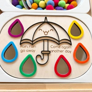 May include: A wooden educational board featuring a central black outline of a smiling umbrella with the text "Rain, rain go away, Come again another day." Surrounding the umbrella are six colorful teardrop-shaped mirrors in purple, yellow, blue, orange, green, and red.