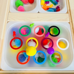 May include: A light-colored wooden table featuring three white plastic bins. The top two bins contain colorful pom-poms and small plastic animal figures. The large bottom bin holds various plastic rings in red, yellow, orange, blue, pink, purple, black, and white, interspersed with multi-colored pom-poms and small plastic animal figures.