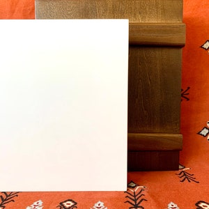 May include: A white rectangular sign with a brown wooden frame. The frame has a raised edge and a decorative groove. The sign is sitting on a red and white patterned fabric.