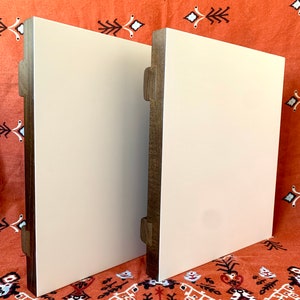 May include: Two square wooden plaques with a white painted surface. The plaques have a dark brown wood frame and are standing upright on a patterned orange fabric.