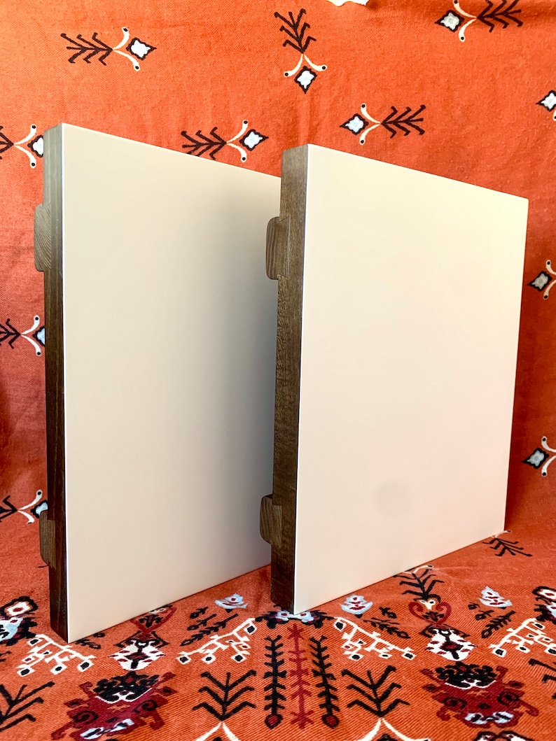May include: Two square wooden plaques with a white painted surface. The plaques have a dark brown wood frame and are standing upright on a patterned orange fabric.
