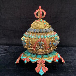 May include: A gold, silver, and turquoise ornate incense burner with a red coral finial. The burner sits on a decorative base with a lotus design.