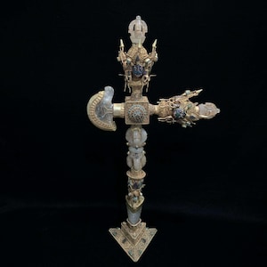 May include: An ornate, gold-colored ritual object with intricate details. The object features a cross-shaped design with a central cylinder and decorative elements, including what appears to be a small axe head and a hand-like structure. The object is adorned with turquoise and other colorful accents.