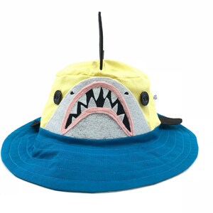May include: A yellow and blue bucket hat with a shark design. The hat features a shark mouth with felt teeth, a gray snout, and a black fin. The brim is blue, and the hat has two black buttons on the sides.