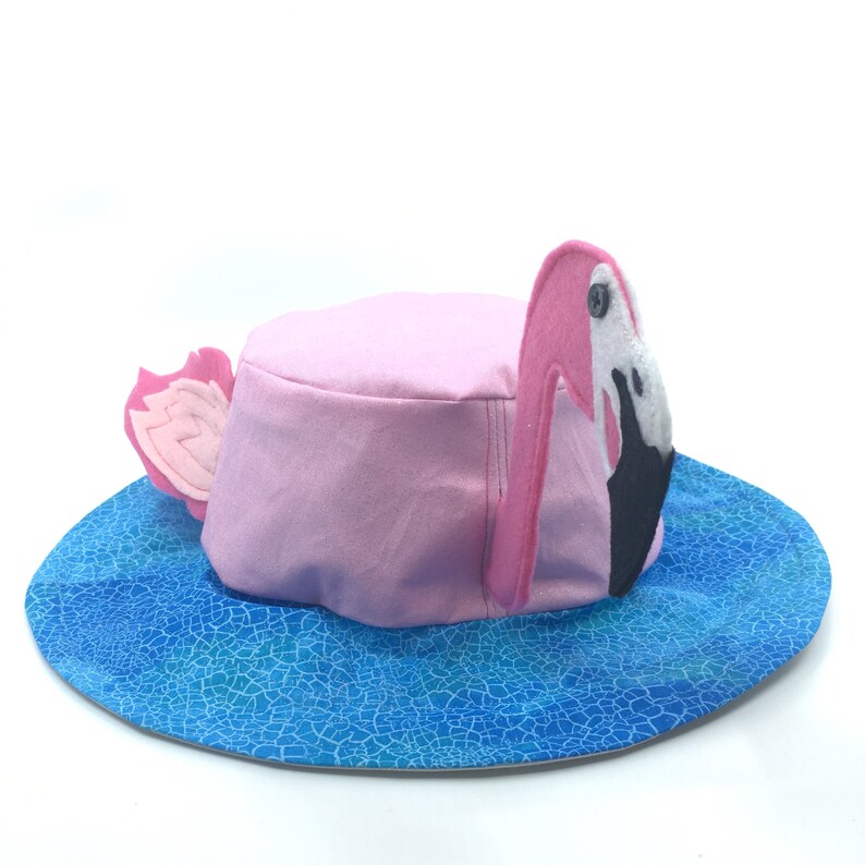 Puede incluir: Sombrero de flamenco rosa con ala azul con estampado de agua. El sombrero tiene una corona rosa y una cabeza de flamenco de fieltro con pico negro y detalles blancos. La cabeza y la cola del flamenco son de fieltro.