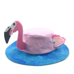 Puede incluir: Un sombrero rosa con forma de flamenco con un ala azul con estampado de agua. El sombrero presenta un cuerpo rosa, una cabeza de flamenco de fieltro con pico negro y alas rosas. El sombrero est&aacute; dise&ntilde;ado para colocarse en la cabeza.