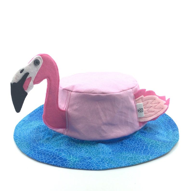 Puede incluir: Un sombrero rosa con forma de flamenco con un ala azul con estampado de agua. El sombrero presenta un cuerpo rosa, una cabeza de flamenco de fieltro con pico negro y alas rosas. El sombrero est&aacute; dise&ntilde;ado para colocarse en la cabeza.