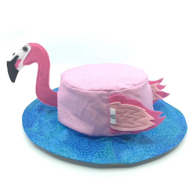 Puede incluir: Un sombrero de flamenco rosa con pico y cabeza de fieltro, y alas de fieltro. El sombrero se asienta sobre un ala azul estampada. La cabeza y el cuello del flamenco se extienden desde el lateral del sombrero. El sombrero es de color rosa claro.