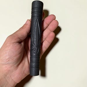 Puede incluir: Un objeto cilíndrico negro con un acabado mate texturizado, que presenta bandas horizontales ranuradas en la parte superior e inferior y un motivo de hoja tallado en el centro. El artículo es alargado y de apariencia sólida, de color gris oscuro.