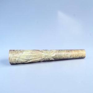 May include: A cylindrical stone pipe featuring intricate hand-carved details. The surface displays a prominent leaf pattern at the centre, surrounded by circular motifs on one end and a smooth, mottled beige and grey texture on the other end.