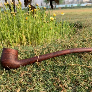Könnte beinhalten: Eine dunkelbraune Tabakpfeife aus Holz mit einem langen, gebogenen Stiel liegt auf grünem Gras. Der Kopf der Pfeife ist strukturiert, und der Stiel verjüngt sich zu einem Mundstück. Der Hintergrund zeigt Gras und Blumen.