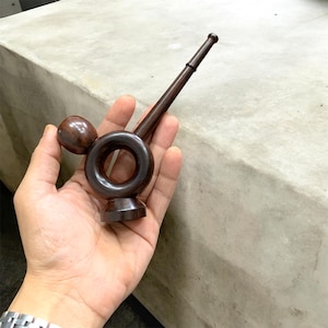 Puede incluir: Una pipa de madera marrón oscuro con una cámara circular y un tallo largo y delgado. La pipa tiene un acabado liso y pulido y se sostiene en la mano de una persona. La pipa está diseñada con un cuenco redondeado y una pequeña esfera decorativa.