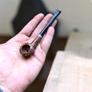 Puede incluir: Una pipa de tabaco de madera con un cuenco marrón oscuro y una boquilla negra. La pipa tiene una banda plateada donde la boquilla se une al cuenco. La pipa se sostiene en una mano.
