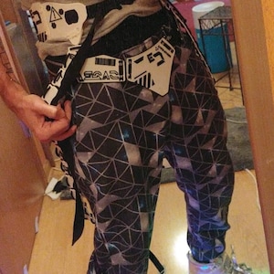 May include: A person wearing black and white geometric print pants with a white belt and silver shoes with light blue LED lights. The shoes have a white strap with the word "GO" printed on it.