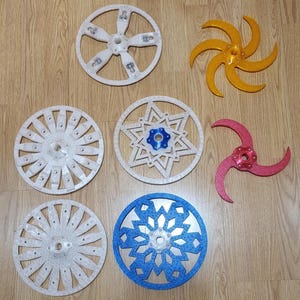 Op de afbeelding: Een verzameling van verschillende geometrische vormen, waaronder wielen en stervormige ontwerpen, in wit, goud, rood en blauw. De objecten zijn gerangschikt op een houten oppervlak, waarbij hun ingewikkelde patronen en vormen worden getoond.