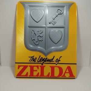 May include: A yellow and gray 3D printed sign with the text "The Legend of ZELDA" in black and red lettering. The sign features a gray shield with a heart, key, and lion design.