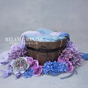Könnte beinhalten: Ein Holzfass gefüllt mit blauem und violettem Stoff, umgeben von violetten und blauen Blumen. Das Fass hat einen schwarzen Metallgriff und ein schwarzes Band um die Oberseite. Der Text "MELANIE MAXINE" und "www.melaniemaxine.com" ist auf dem Fass sichtbar.