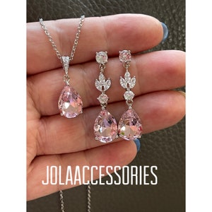 Könnte beinhalten: Ein zartes Braut-Schmuckset mit einem birnenförmigen rosa Kristall-Anhänger und passenden Ohrringen mit floralen Akzenten. Die Halskette hat eine silberne Kette und die Ohrringe haben silberne Ohrstecker.