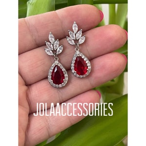 May include: A pair of silver dangle earrings featuring a red teardrop-shaped stone surrounded by clear rhinestones. The earrings have a leaf-shaped design with clear rhinestones.