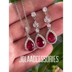 May include: A silver necklace and earrings set with a teardrop-shaped red gemstone surrounded by clear cubic zirconia stones. The pendant and earrings feature a delicate floral design.