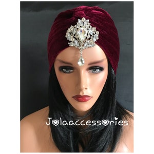 May include: A burgundy velvet turban with a silver and crystal jeweled headpiece. The headpiece has a teardrop-shaped crystal pendant.