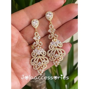 Peut inclure: Une paire de boucles d'oreilles pendantes dorées avec des cristaux clairs et étincelants. Les boucles d'oreilles présentent un cristal en forme de goutte d'eau en haut, avec des motifs floraux et géométriques complexes. Le texte "Jola accessories" est visible.
