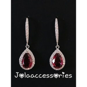 May include: A pair of silver dangle earrings with a teardrop-shaped red gemstone surrounded by clear cubic zirconia stones. The earrings are on a black background.