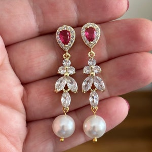Peut inclure: Boucles d'oreilles pendantes dorées avec une pierre rouge en forme de poire, des zircons cubiques transparents et une perle blanche au bas.