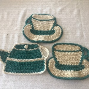 Puede incluir: Juego de té de ganchillo hecho a mano, que incluye una tetera y dos tazas con platillos. El conjunto es de color turquesa y crema, con un diseño a rayas. La tetera tiene un botón y asas. Sobre un fondo blanco.