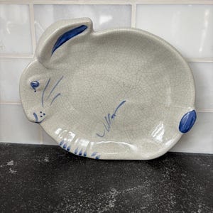 Könnte beinhalten: Ein keramikförmiger Teller in Hasenform mit einer rissigen, cremefarbenen Glasur. Der Teller hat blaue Akzente an Ohren, Augen, Nase und Schwanz. Der Teller steht auf einer schwarzen Oberfläche.