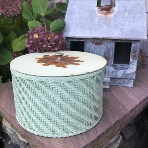 May include: A light green wicker basket with a white lid and a metal knob. The lid has a brown and orange design. The basket is sitting on a stone surface.